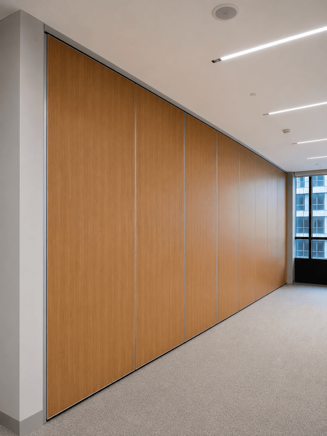 Modern office space featuring a large wood-paneled movable partition wall, grey carpet, and linear ceiling lights.